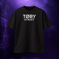 Camiseta negra de manga corta TØBY STREET con logotipo gráfico blanco en el pecho sobre fondo urbano morado